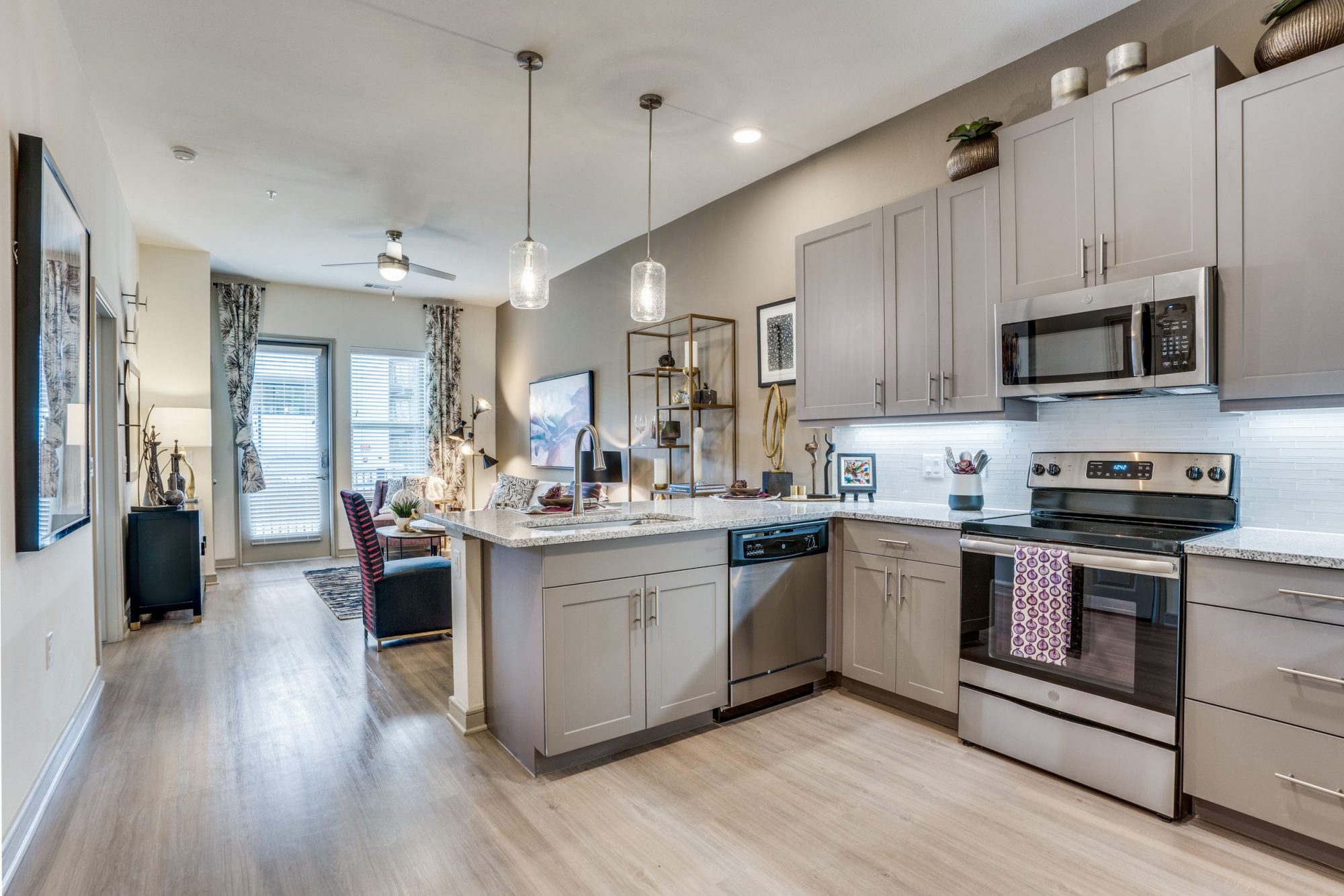 View from the L shaped kitchen with marble countertops, stainless steel appliances, wood look floors, upper and lower cabinets, pendant lighting and a view into the living/dining room