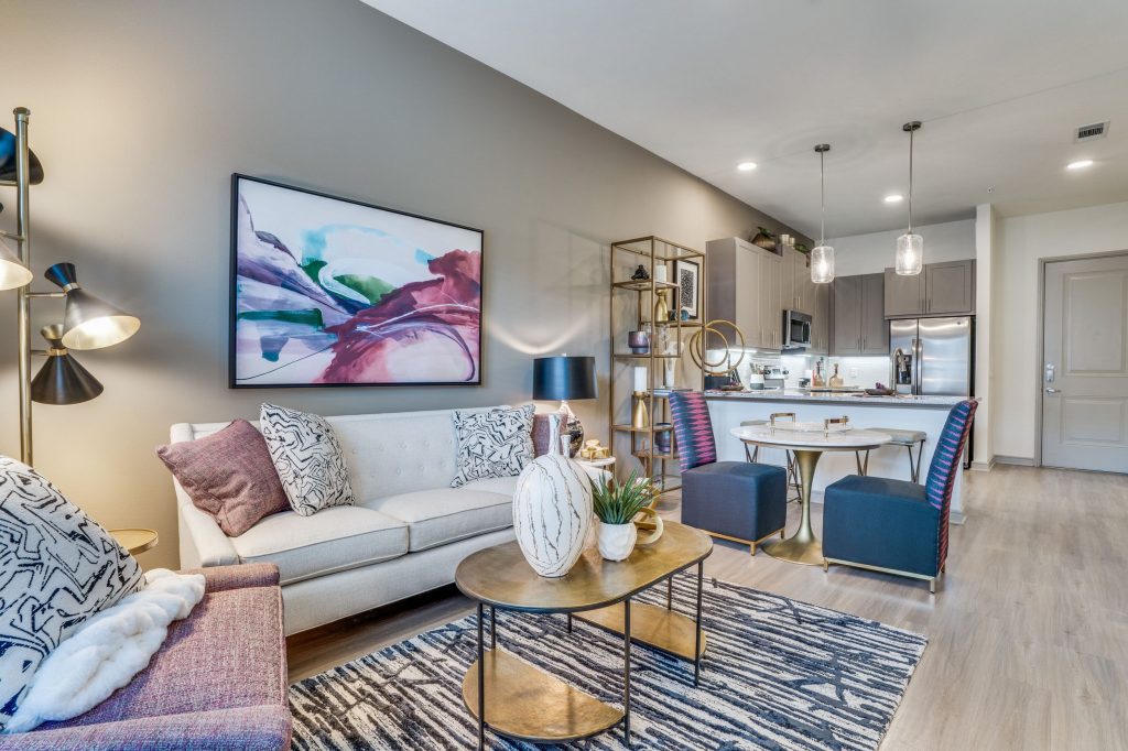 Living room with striped pattern rug, coffee table, an armchair and couch with pillows, abstract artwork on the wall and a 2 seater dining table. Kitchen is in the background