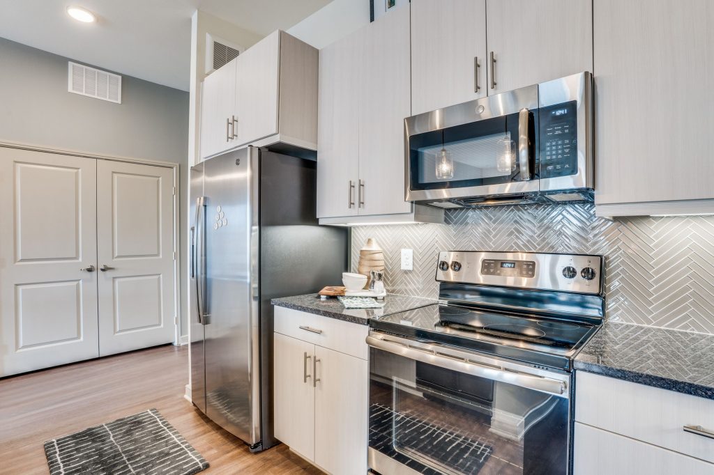 Kitchen with stainless steel appliances, upper and lower cabinets, custom gray tile backsplash, wood look floors and double doors in the background
