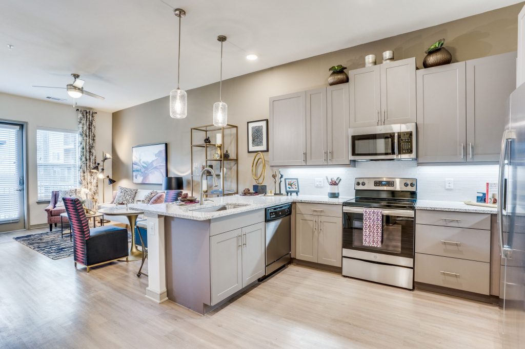 View from the L shaped kitchen with marble countertops, stainless steel appliances, wood look floors, upper and lower cabinets, pendant lighting and a view into the living/dining room.