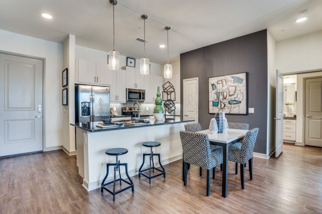 Interior shot of the kitchen with pendant lighting, bar stool seating, stainless steel appliances, and a 4 seater dining table. Kitchen has white cabinets, wood look floors and a gray accent wall