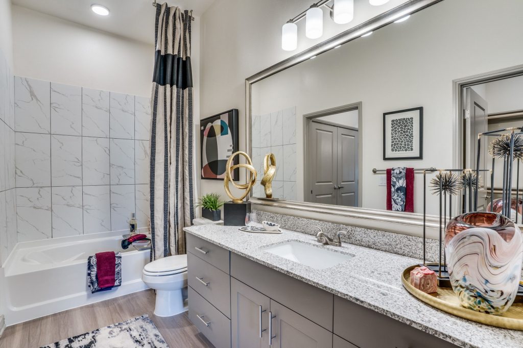 Bathroom shot with a marble countertop single sink vanity, large framed mirror, wood look floors and a full size shower tub combo with tile backsplash.