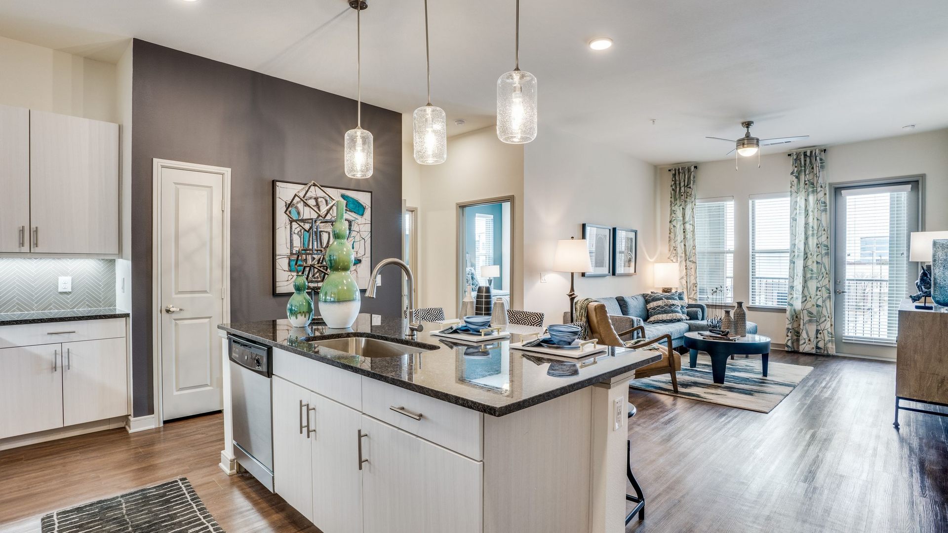 View from the kitchen with marble top island with dishwasher, sink and pendant lighting. Pantry door is on the left. Living room is in the background with a couch, coffee table, and wood look floors