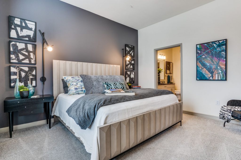 Bedroom with carpet, queen size bed with decorative pillows, a gray accent wall, abstract artwork hanging and black nightstands. Doorway on the right opens to the bathroom. Photo 6: Bedroom with carpet and teal accent wall, a full size bed, large window with patterned drapes and dual side tables with lamps