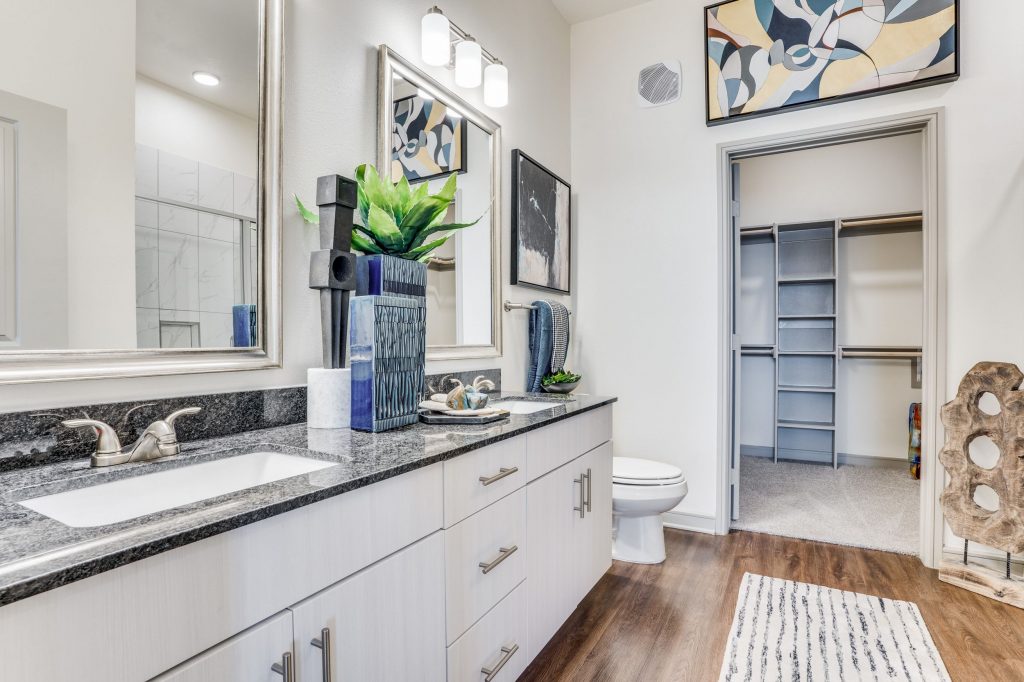 Bathroom with gray marble countertop, dual sink vanity with individual framed mirrors, white cabinets, wood look floors, and walk in closet with shelving on the right.