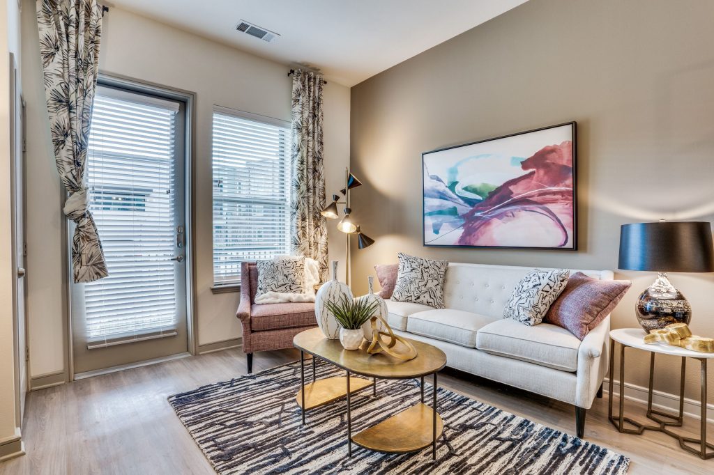 Living room with taupe accent wall, couch with pillows, coffee table and comfortable armchair. A patterned rug sits below the table with wood look floors and a window and window door looking to the patio