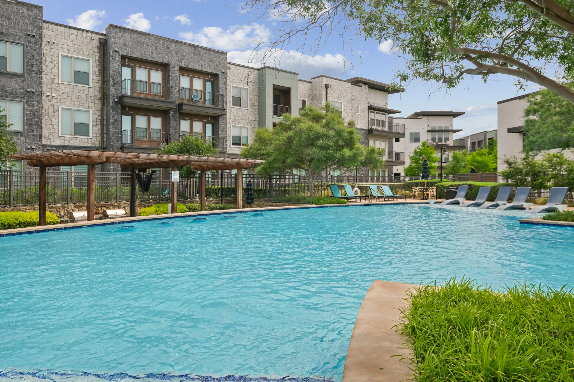 A modern apartment complex with stone and gray buildings overlooks a large outdoor swimming pool, surrounded by lounge chairs, greenery, and a wooden pergola under a partly cloudy sky.